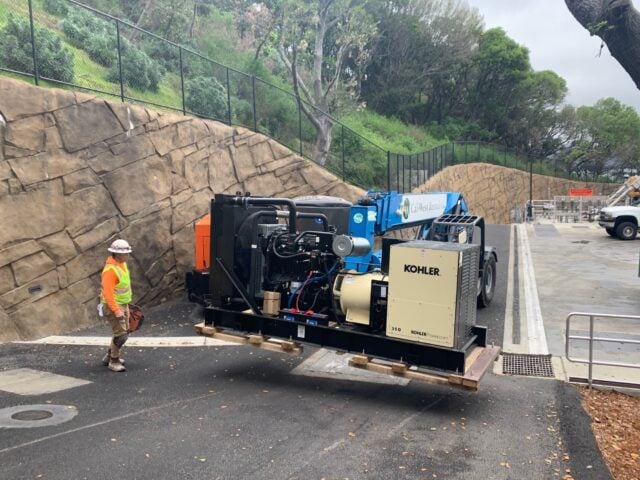 A backup electrical generator being transported by an industrial forklift as a worker watches on.