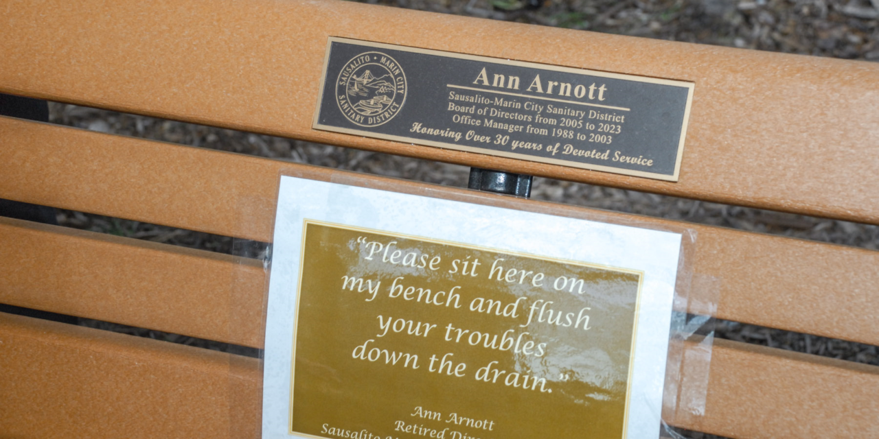 Park bench, honoring Ann Arnott, showing a plaque that reads "Honoring Over 30 years of Devoted Service" and a paper taped to the bench that reads "Please sit here on my bench and flush your troubles down the drain."